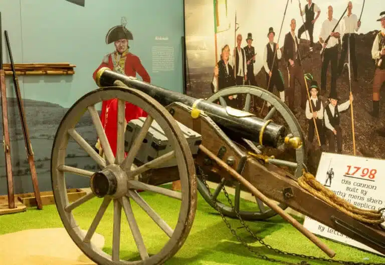 Display at the National 1798 Rebellion Centre, Enniscorthy, showing a replica cannon on wooden wheels, surrounded by muskets, a figure in red uniform and a mural of reenactors dressed as 1798 rebels.