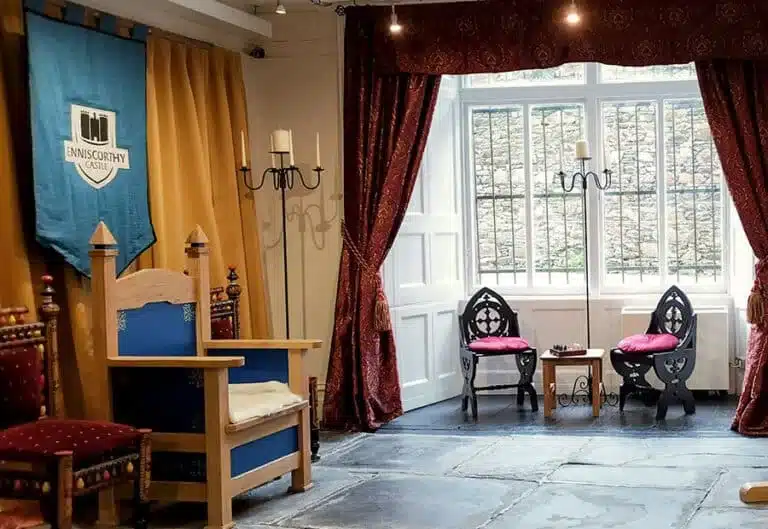 Interior room in Enniscorthy Castle featuring a medieval-style throne, decorative chairs, banners and large curtained windows.