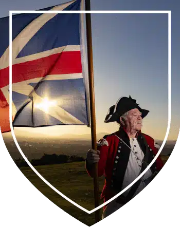 Man dressed in a red 18th-century military uniform holding a large British union flag at sunset.