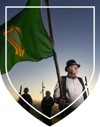 Man in period clothing holding a green flag with a gold harp emblem representing Irish rebels, standing with others at dusk.